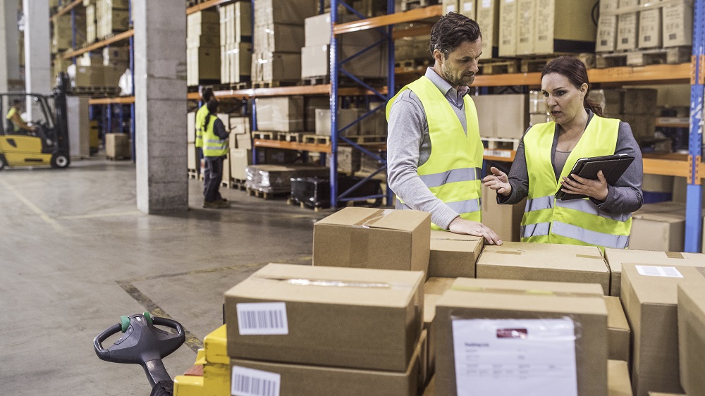 Displeased woman holding a digital tablet pointing at cardboard boxes and giving instructions to an employee in a warehouse. Forklift and other workers visible in the background.